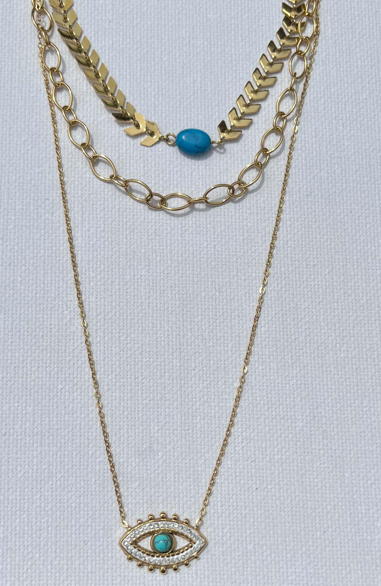 Découvre notre collier multi-chaînes, en acier inoxydable et plaqué or. Ce bijou se compose de trois chaînes, chacune apportant sa touche unique. La première chaîne est une élégante maille chevron, ornée d'une perle turquoise. La seconde chaîne, plus courte, présente des maillons ovales entrelacés, créant un jeu de textures. Enfin, la troisième chaîne, plus fine et minimaliste, met en avant un pendentif en forme d’œil, serti de zircons et d'une pierre turquoise centrale.