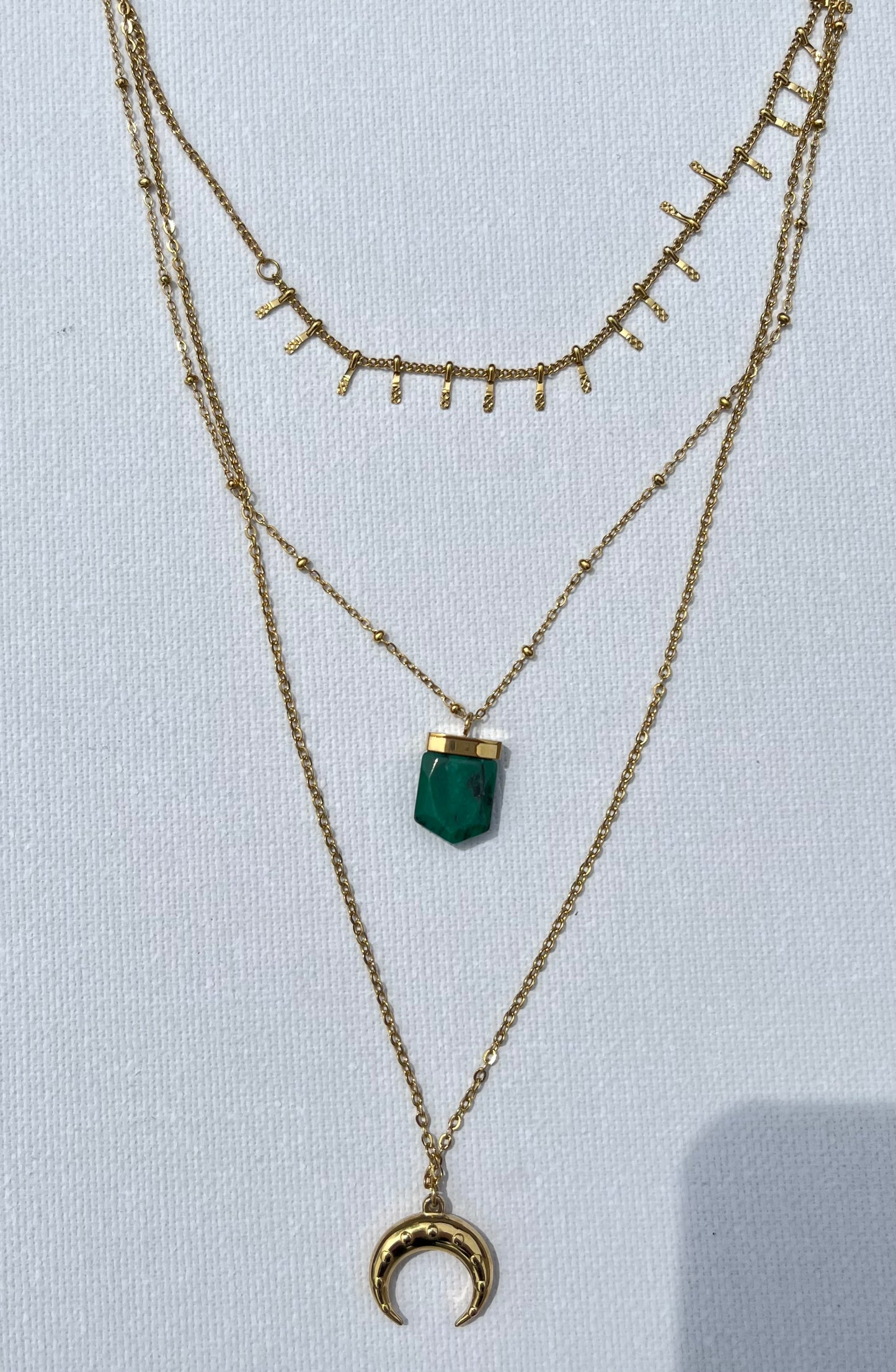 Découvre ce sublime collier multi-rangs, en acier inoxydable et plaqué or ! Composé de trois chaînes distinctes, il est à la fois délicat et audacieux. La première chaîne, ornée de petites barres texturées, apporte une touche sophistiquée. La seconde, plus fine, est sublimée par un pendentif en pierre de malachite, taillé en pointe. Enfin, la troisième chaîne, légèrement plus longue, se termine par un pendentif en forme de croissant de lune, symbole de féminité.