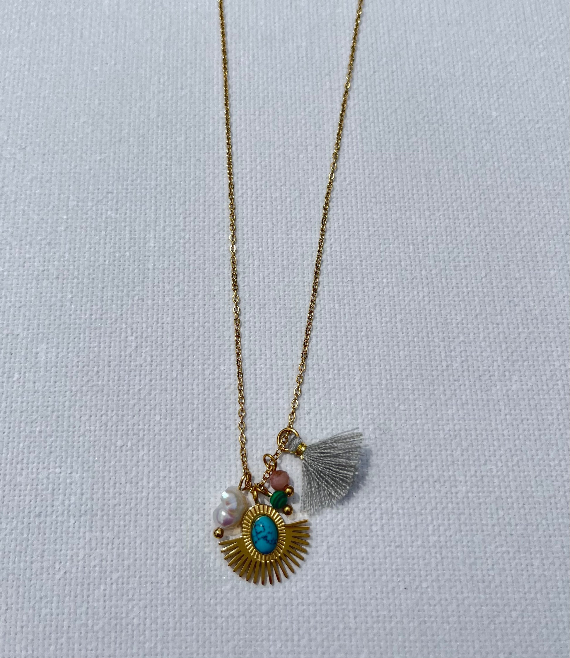 Opte pour ce collier, en acier inoxydable et plaqué or, parfait pour sublimer tes tenues. Sa chaîne fine en maille forçat met en valeur un pendentif médaillon solaire, rehaussé d'une pierre turquoise au centre, symbole de protection et de sérénité. Il est accompagné d'une perle nacrée, d'une pierre rose pâle et d’un pompon gris délicat. Ce collier polyvalent se marie aussi bien avec un look décontracté qu’avec une tenue plus sophistiquée.