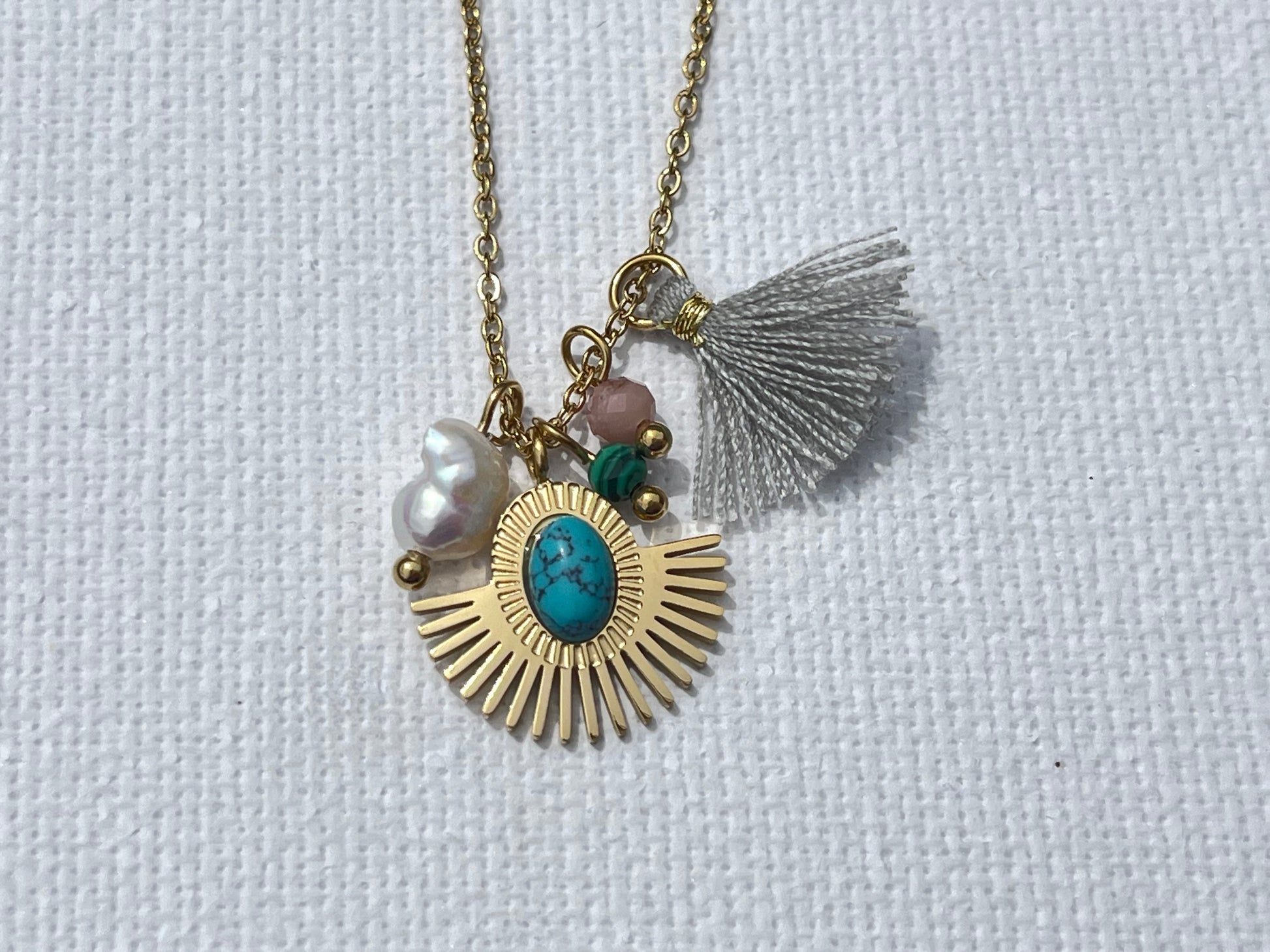 Opte pour ce collier, en acier inoxydable et plaqué or, parfait pour sublimer tes tenues. Sa chaîne fine en maille forçat met en valeur un pendentif médaillon solaire, rehaussé d'une pierre turquoise au centre, symbole de protection et de sérénité. Il est accompagné d'une perle nacrée, d'une pierre rose pâle et d’un pompon gris délicat. Ce collier polyvalent se marie aussi bien avec un look décontracté qu’avec une tenue plus sophistiquée.